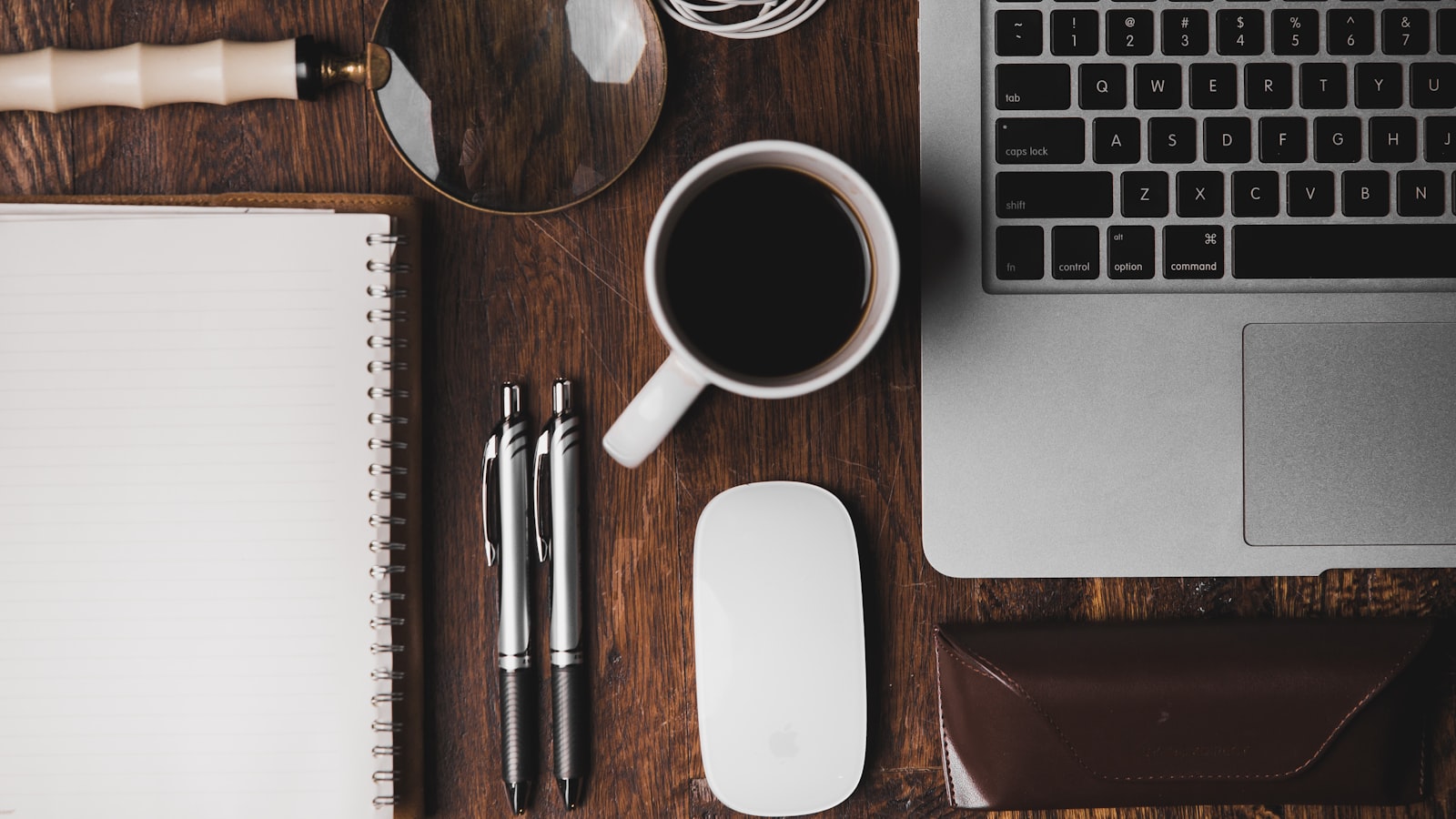 A notebook and pen on a wooden table beside a cup of coffee.